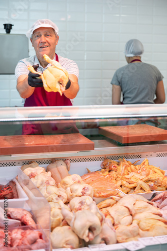 Skilled senior man, owner of butcher shop preparing meat products for sale in glass refrigerated showcase, offering fresh raw chickens