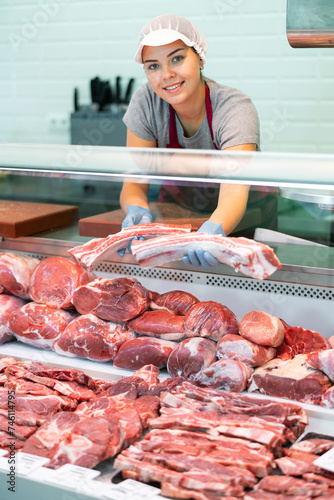 Smiling skilled young female butcher working behind counter in butchery, showing cuts of fresh raw veal short ribs