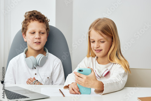 Happy children studying at home using laptops and tablets for elearning, sitting at a table in their cozy living room The boy and girl, fully concentrated, are connected to the internet, wearing