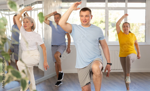 Positive middle-aged male instructor leading energetic zumba class for group of seniors in mirrored fitness studio..