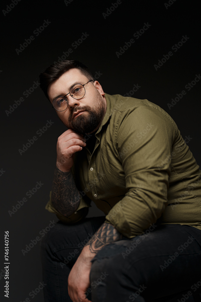 A contemplative man in an olive shirt rests his chin on his hand, his tattoos adding character to the portrait
