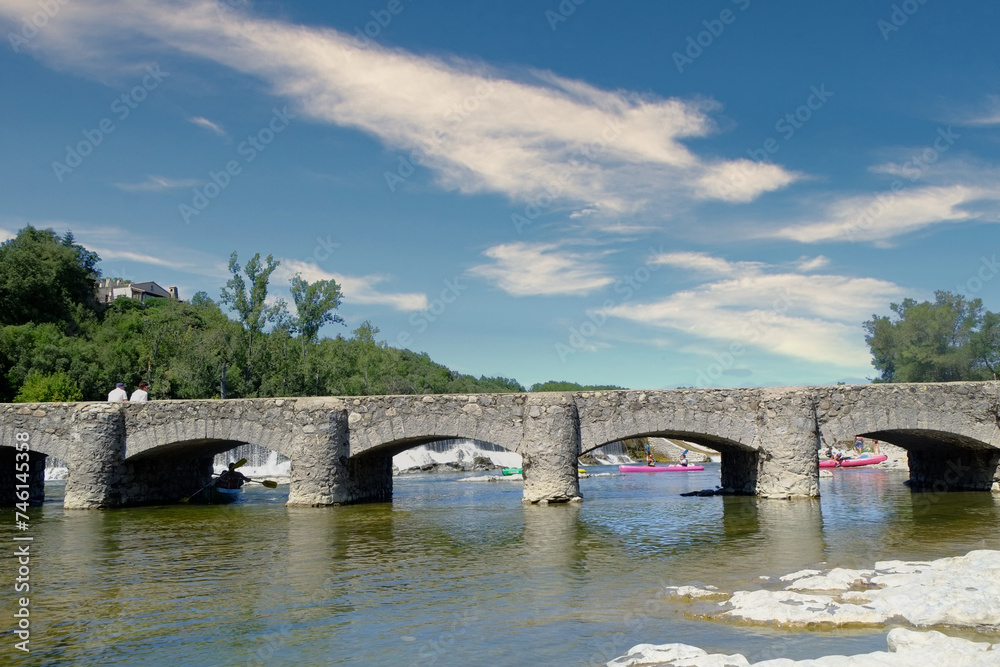 This vibrant and sunny scene captures the essence of summer in the Ardeche region, with individuals leisurely canoeing under a majestic old stone bridge that crosses the calm river. The bridge, with This vibrant and sunny scene captures the essence of summer in the Ardeche region, with individuals leisurely canoeing under a majestic old stone bridge that crosses the calm river. The bridge, with