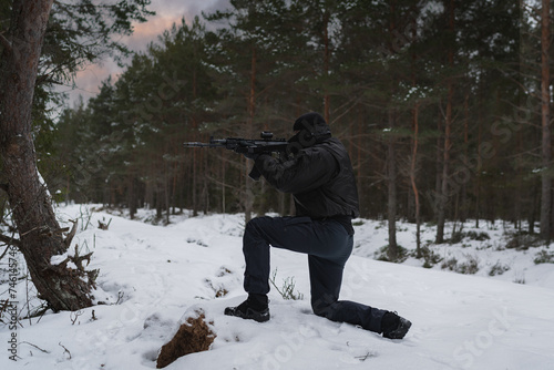 A military man with a modern assault rifle takes aim while sitting on his knee in the winter forest.