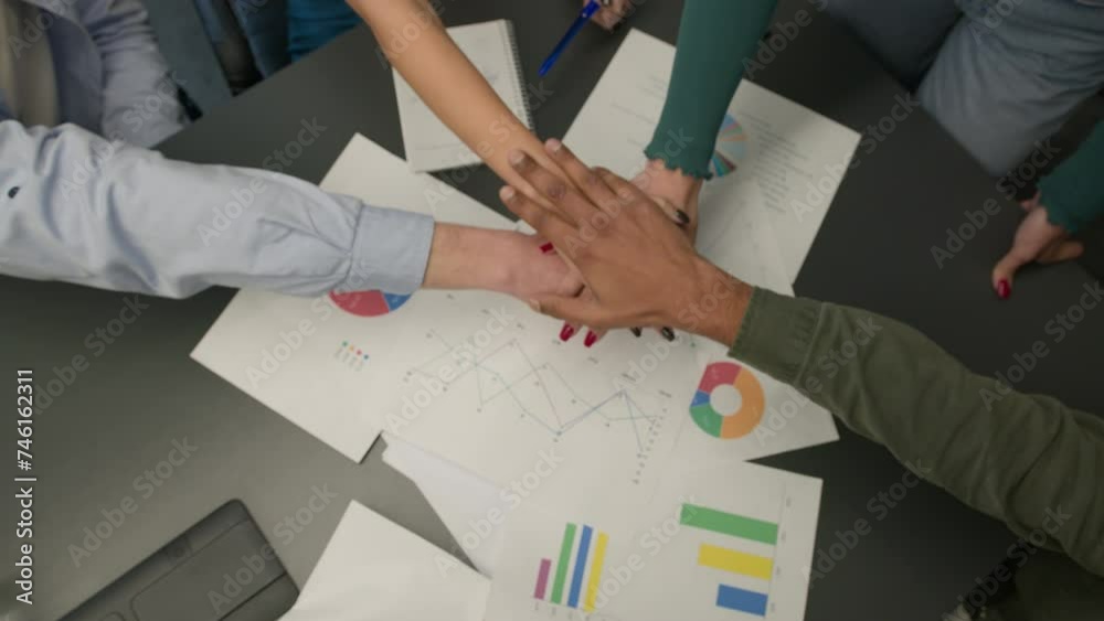 Diverse people women men stacking hands together in pile unite gesture on table with project paperwork group students team co-workers united stack palms connected in teamwork unity close up top view
