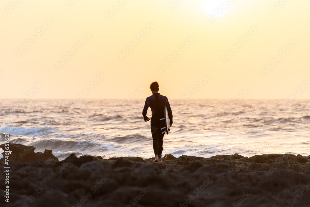 Rear View of Young Surfer going to the water Against a Orange Sunset Sky in Canary Island.Copy Space Rear View of Young Surfer going to the water Against a Orange Sunset Sky in Canary Island.Copy Space