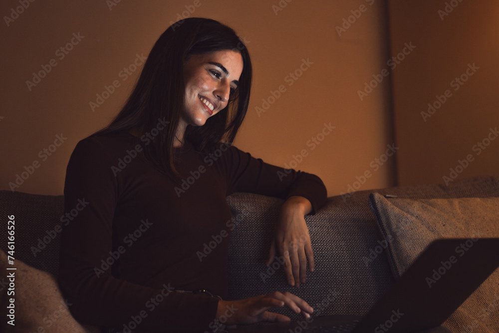 Close up of one young happy cheerful woman smiling and using laptop computer at home sitting on sofa working and studying alone at late night. Millennial girl surfing the net indoor with light screen