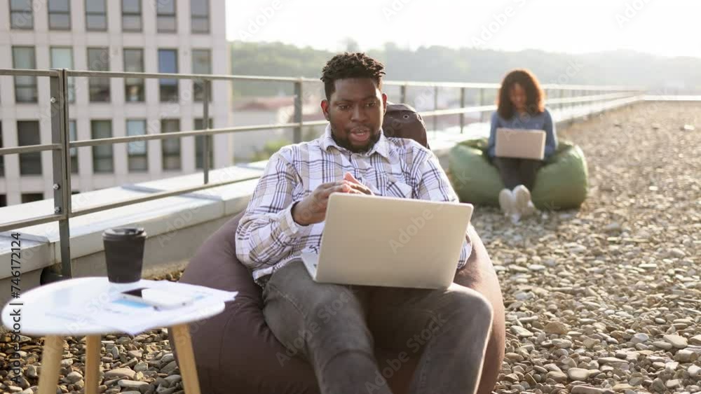 Happy African American male person in casual clothing gesture at laptop camera on flat roof of building, while his female colleague working in blurred background. Happy African American male person in casual clothing gesture at laptop camera on flat roof of building, while his female colleague working in blurred background.