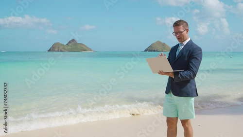 Young caucasian gentleman in glasses teleconferencing on laptop standing on beach surrounded by the scenic view of crystal clear water of ocean, blue sky. High quality 4k footage photo