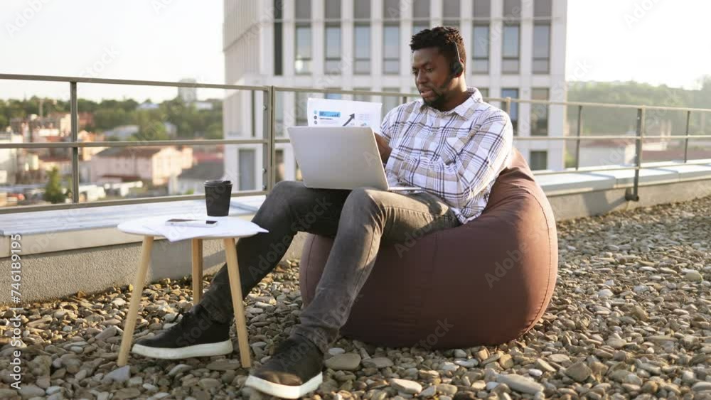 Young African male in casual clothes giving online presentation using headset and computer outside office. Remote manager brainstorming about statistics shown by infographics on terrace. Young African male in casual clothes giving online presentation using headset and computer outside office. Remote manager brainstorming about statistics shown by infographics on terrace.