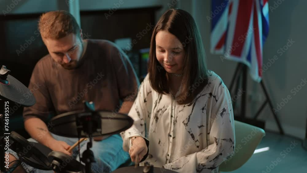 Slow motion. Under the guidance of a teacher, a girl drummer plays with drumsticks on an electronic drum kit, sitting in the studio of a music school Slow motion. Under the guidance of a teacher, a girl drummer plays with drumsticks on an electronic drum kit, sitting in the studio of a music school
