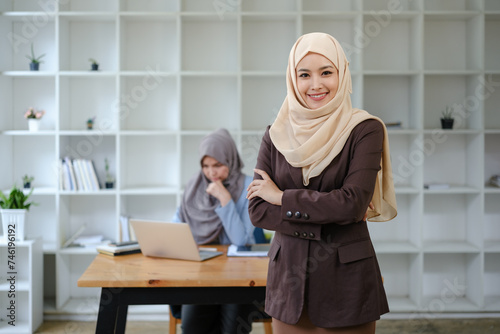 Beautiful young working woman in hijab standing in office, smiling. Portrait of confident muslim businesswoman. Modern office with big window, friend working at desk on background. 