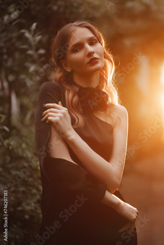 Caucasian woman in black dress, sitting outdoors during golden hour. Her wavy hair is highlighted by the sun. Concept for fashion advertisements or storytelling.
