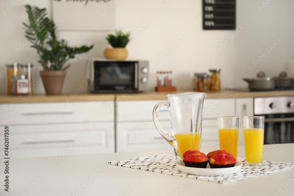 Jug and glass of fresh orange juice with muffins on table in kitchen Jug and glass of fresh orange juice with muffins on table in kitchen