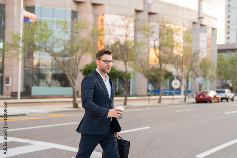Businessman with backpack walking in city. Side view of male entrepreneur in classy suit and with takeaway drink walking along crosswalk while commuting to work in morning. businessman in the street Businessman with backpack walking in city. Side view of male entrepreneur in classy suit and with takeaway drink walking along crosswalk while commuting to work in morning. businessman in the street