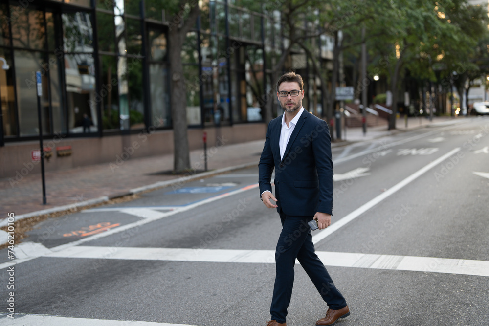 Businessman with backpack walking in city. Side view of male entrepreneur in classy suit and with takeaway drink walking along crosswalk while commuting to work in morning. businessman in the street Businessman with backpack walking in city. Side view of male entrepreneur in classy suit and with takeaway drink walking along crosswalk while commuting to work in morning. businessman in the street