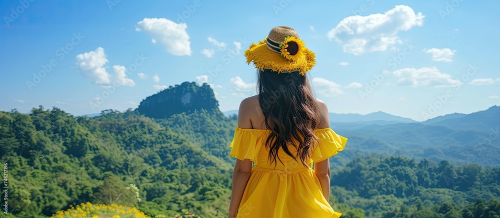 A young woman from Thailand, dressed in a yellow dress and sunflower hat, gazes at the majestic green mountains under a clear sky.