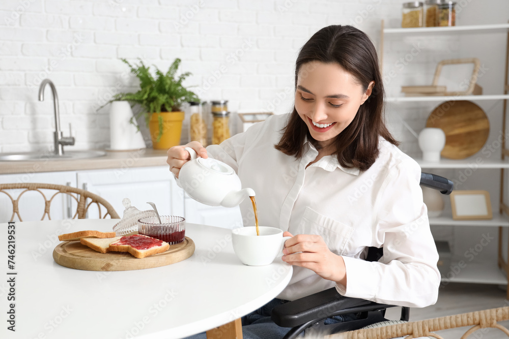 Happy young woman in wheelchair pouring tea into cup at home Happy young woman in wheelchair pouring tea into cup at home