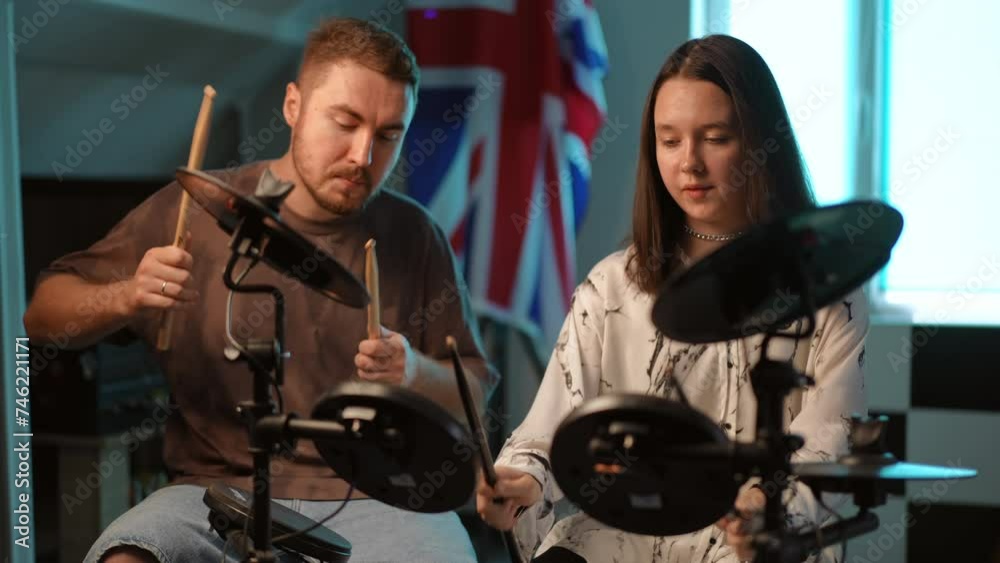 A male teacher shows an aspiring drummer girl how to hit an electronic drum kit with drumsticks. The girl, following the teacher, tries to repeat complex movements, sitting in the music school A male teacher shows an aspiring drummer girl how to hit an electronic drum kit with drumsticks. The girl, following the teacher, tries to repeat complex movements, sitting in the music school
