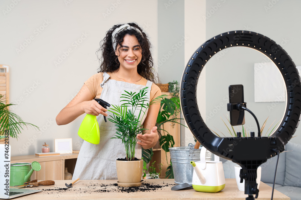 Obraz premium Female African-American gardener with plant and spray water bottle recording video at home