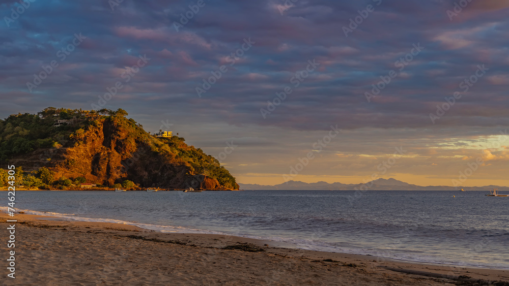 Obraz premium Sunset on a tropical island. The ocean waves are foaming on the sandy beach. The clouds in the sky are highlighted in pink. A hill in the distance. Mountains on the horizon. The golden hour. Nosy Be