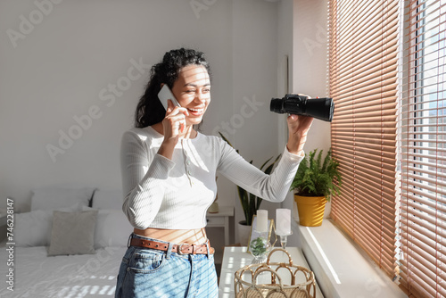 Young African-American woman with binoculars talking by mobile phone near window at home photo