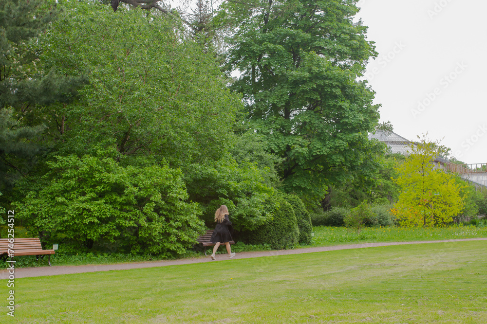 Young woman walking in green spring nature park. Distance view of girl in black dress and hat with trees on background Young woman walking in green spring nature park. Distance view of girl in black dress and hat with trees on background