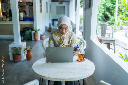A beautiful, charismatic Asian woman wearing a casual hijab is working or doing assignments in a cafe. Seen using a laptop. The concept of working anywhere. photo