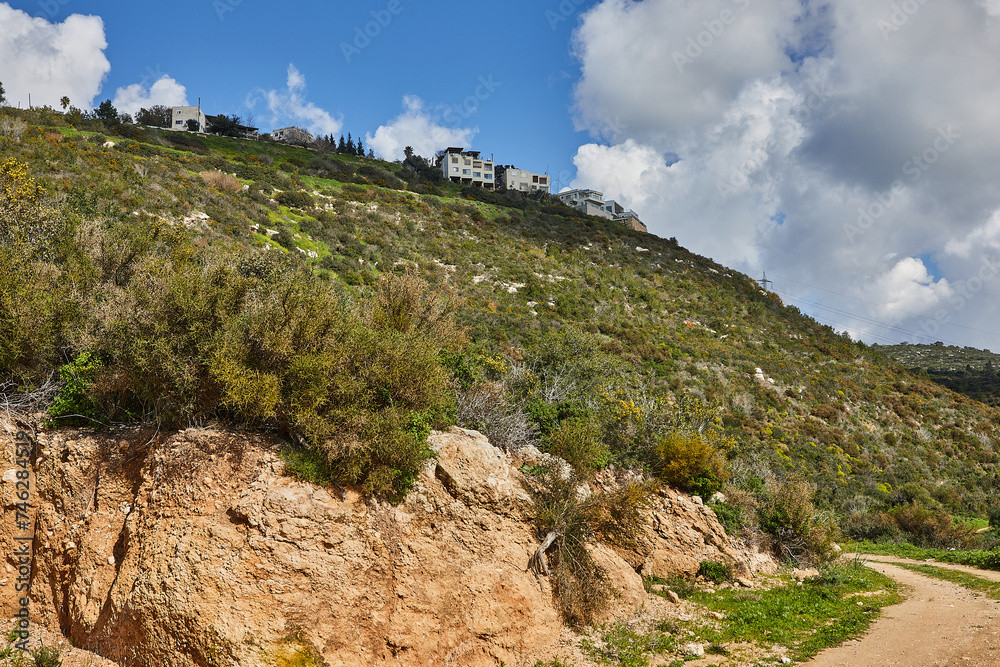Rocky hill with a dirt road in front under a blue sky with white clouds and a modern villa on top