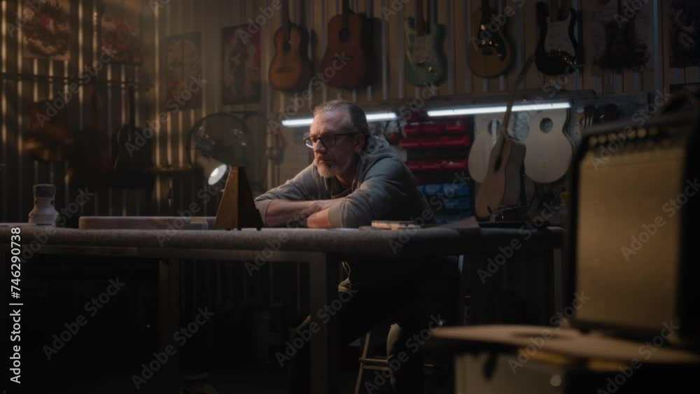 Tired male artisan takes off glasses, looks at metronome. Mature carpenter relaxes after long workday in modern workshop for making musical instruments. Handcrafted wooden guitars hang on the wall. Tired male artisan takes off glasses, looks at metronome. Mature carpenter relaxes after long workday in modern workshop for making musical instruments. Handcrafted wooden guitars hang on the wall.