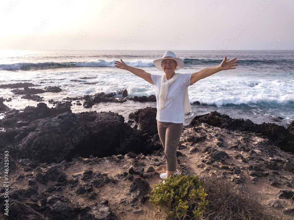 Smiling senior woman on a rocky beach with open arms  looking at camera while ocean waves crashing with white foam. Concept of relaxed elderly person enjoying retirement lifestyle