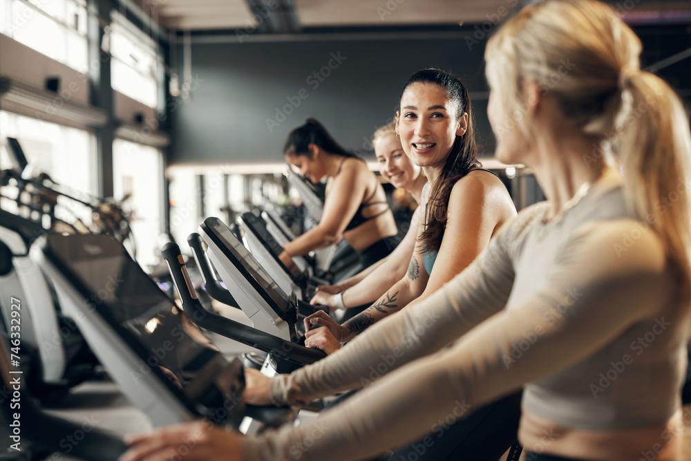 Smiling young woman and a group of female friends in sportswear riding on stationary bikes during a cardio workout class together at the gym Smiling young woman and a group of female friends in sportswear riding on stationary bikes during a cardio workout class together at the gym