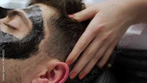 Barber applies black charcoal mask on clients face in shop. Man relaxes during skincare routine. Grooming ritual, pore cleansing, detox treatment. Male pampering for clear skin, facial care.