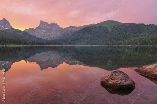 Ideal pink sunset on the banks of the lake in the mountains scenery. Stones in the water, coniferous forest, rocky cliff and mirror reflection in clean water. Ergaki, Siberia Russia Krasnoyarsk region photo