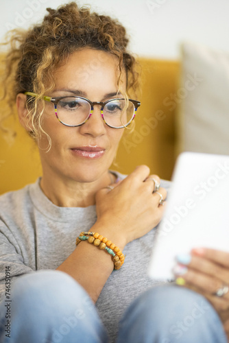 Front view of woman using tablet at home in relaxation lifestyle activity. Cute female people alone sitting against yellow sofa in indoor leisure technology connection activity. Modern lady eyewear photo