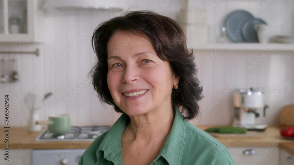 portrait of elderly cheerful woman with dark hair standing in cozy kitchen and smiling portrait of elderly cheerful woman with dark hair standing in cozy kitchen and smiling