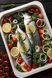 Raw fish with lemon in baking dish and vegetables on black wooden table, flat lay