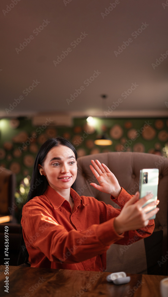 Smiling hipster girl waving hand hello during online video call via cellphone, sitting in cafe. Attractive business woman or student having conference in internet via smartphone. Copy space Smiling hipster girl waving hand hello during online video call via cellphone, sitting in cafe. Attractive business woman or student having conference in internet via smartphone. Copy space