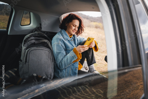Young happy woman sitting in an open car trunk taking pictures with a camera. Traveling by car, communication in travel concept © olezzo