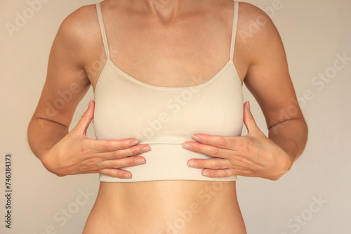 A young woman in beige underwear holds her small breasts with both hands from below. Conducts a breast self-examination. Isolated on a beige background. Women's health