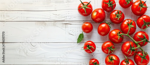 A top view of a collection of vibrant red tomatoes scattered on a clean, white wooden table. The tomatoes appear fresh and ripe, with green stems attached. The setting provides ample space for