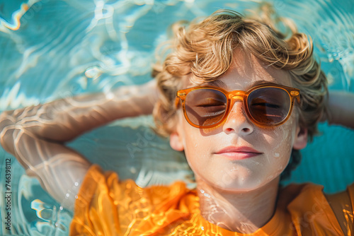 Top view of a cheerful curly-haired blond boy in a T-shirt and sunglasses lying in the pool with his hands behind his head.