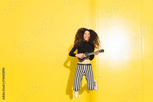 Smiling woman playing ukulele and jumping against yellow background photo