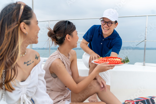 Asian man waiter serving fresh fruit to woman passenger tourist on luxury catamaran boat yacht sailing in the ocean. Attractive girl enjoy outdoor lifestyle travel on summer holiday beach vacation.