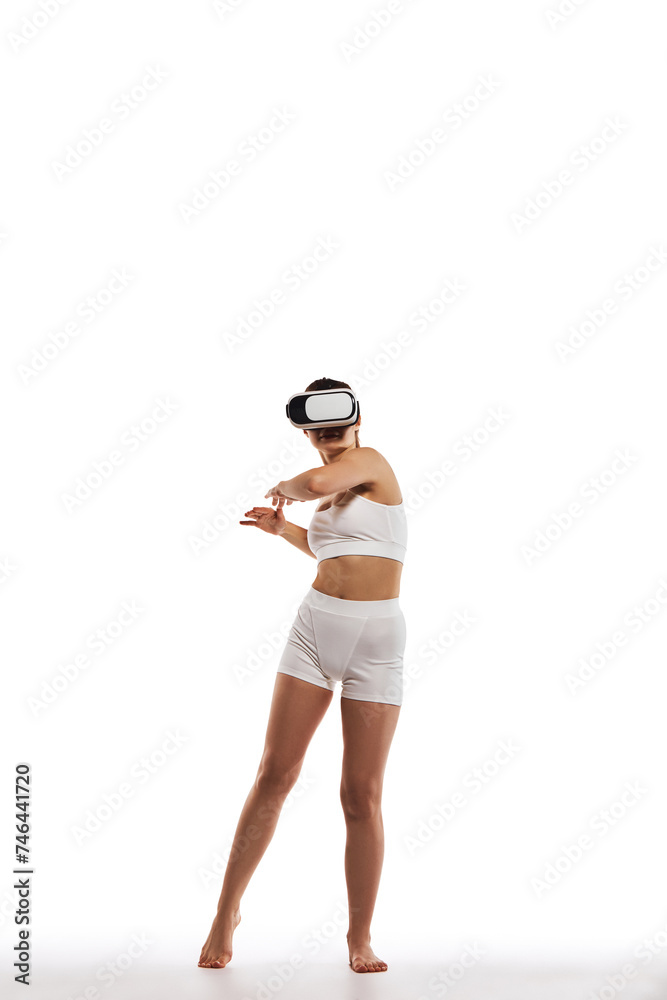 Keeping body in tone. Full-length image of young girl with slim body, wearing VR glasses and training against white studio background. Concept of body and health care, sport, female beauty, wellness Keeping body in tone. Full-length image of young girl with slim body, wearing VR glasses and training against white studio background. Concept of body and health care, sport, female beauty, wellness