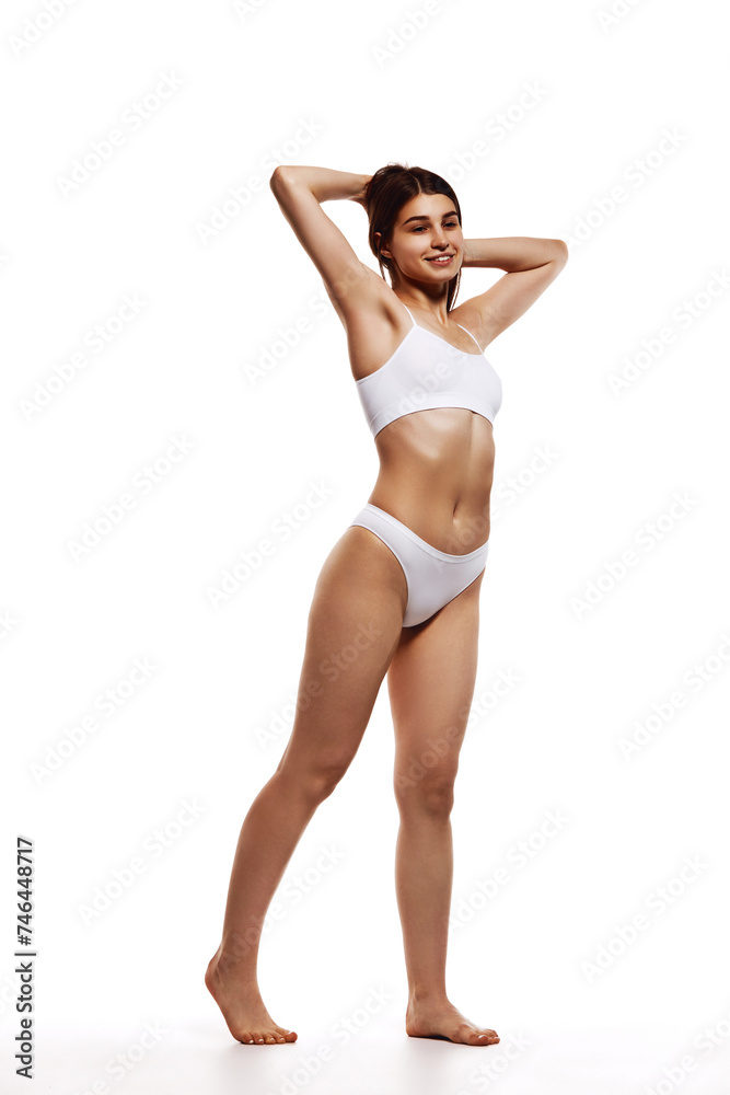 Full-length image of beautiful young girl with tanned, slim, fit body posing in underwear against white studio background. Concept of body and health care, sport, female beauty, wellness Full-length image of beautiful young girl with tanned, slim, fit body posing in underwear against white studio background. Concept of body and health care, sport, female beauty, wellness