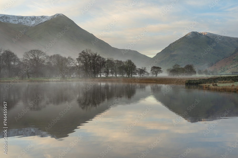 Misty calm morning with trees on lake shoreline and mountains in background at Brothers Water in The English Lake District.