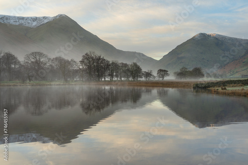 Misty calm morning with trees on lake shoreline and mountains in background at Brothers Water in The English Lake District.