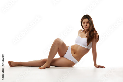 Beautiful young woman with slim fit body posing on floor in comfortable cotton underwear against white studio background. Concept of body and health care, sport, female beauty, wellness