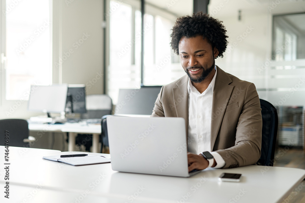 Confident and approachable African-American businessman sits at his desk in a modern office, using a laptop. Concept of strategic thinking in a professional business atmosphere Confident and approachable African-American businessman sits at his desk in a modern office, using a laptop. Concept of strategic thinking in a professional business atmosphere