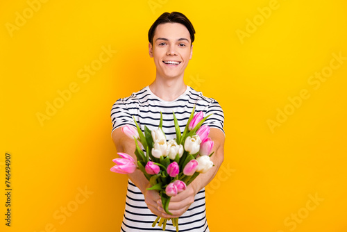 Photo of positive student dressed striped t-shirt presenting you bouquet of flowers on valentine day isolated on yellow color background